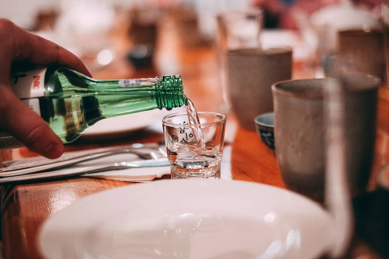 飲食店の木のテーブルで、緑の瓶から小さなグラスに焼酎を注ぐ手
