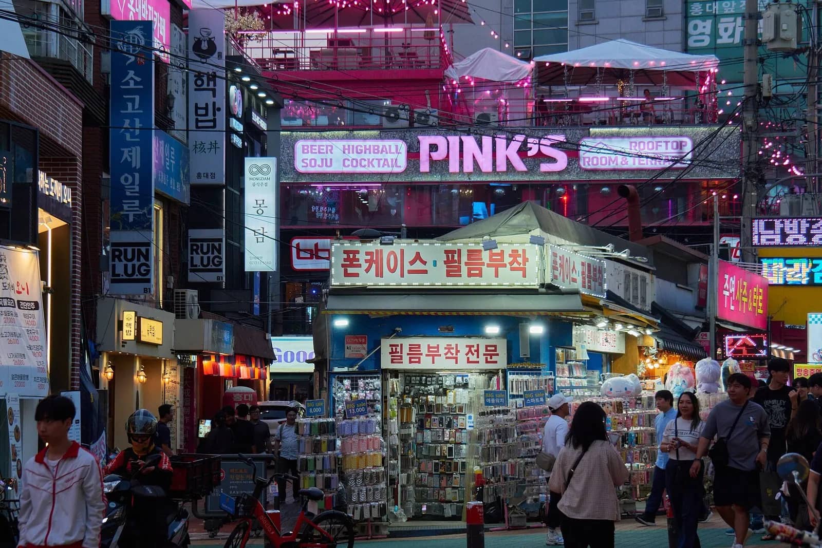 傍晚首爾弘大熱鬧的街角，上方有酒吧霓虹招牌和屋頂露台