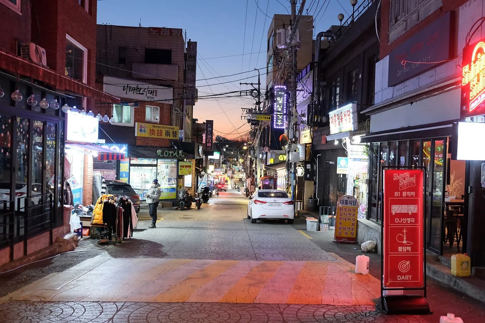 Side street in Itaewon at dusk with neon bar signs, small shops and a parked car