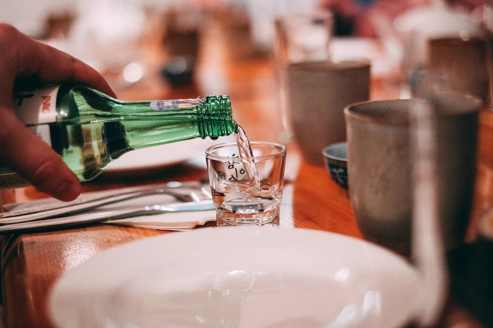 Hand pouring soju from a green bottle into a small glass on a wooden restaurant table