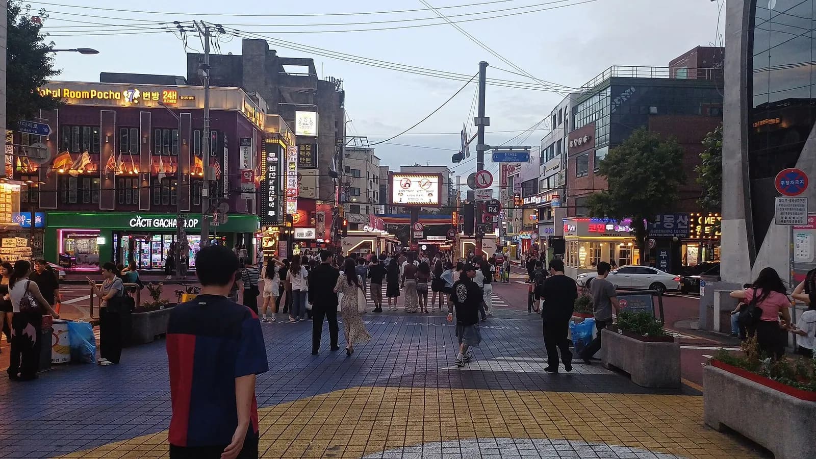 People seen from behind cross a tiled Hongdae plaza toward lit shopfronts at dusk