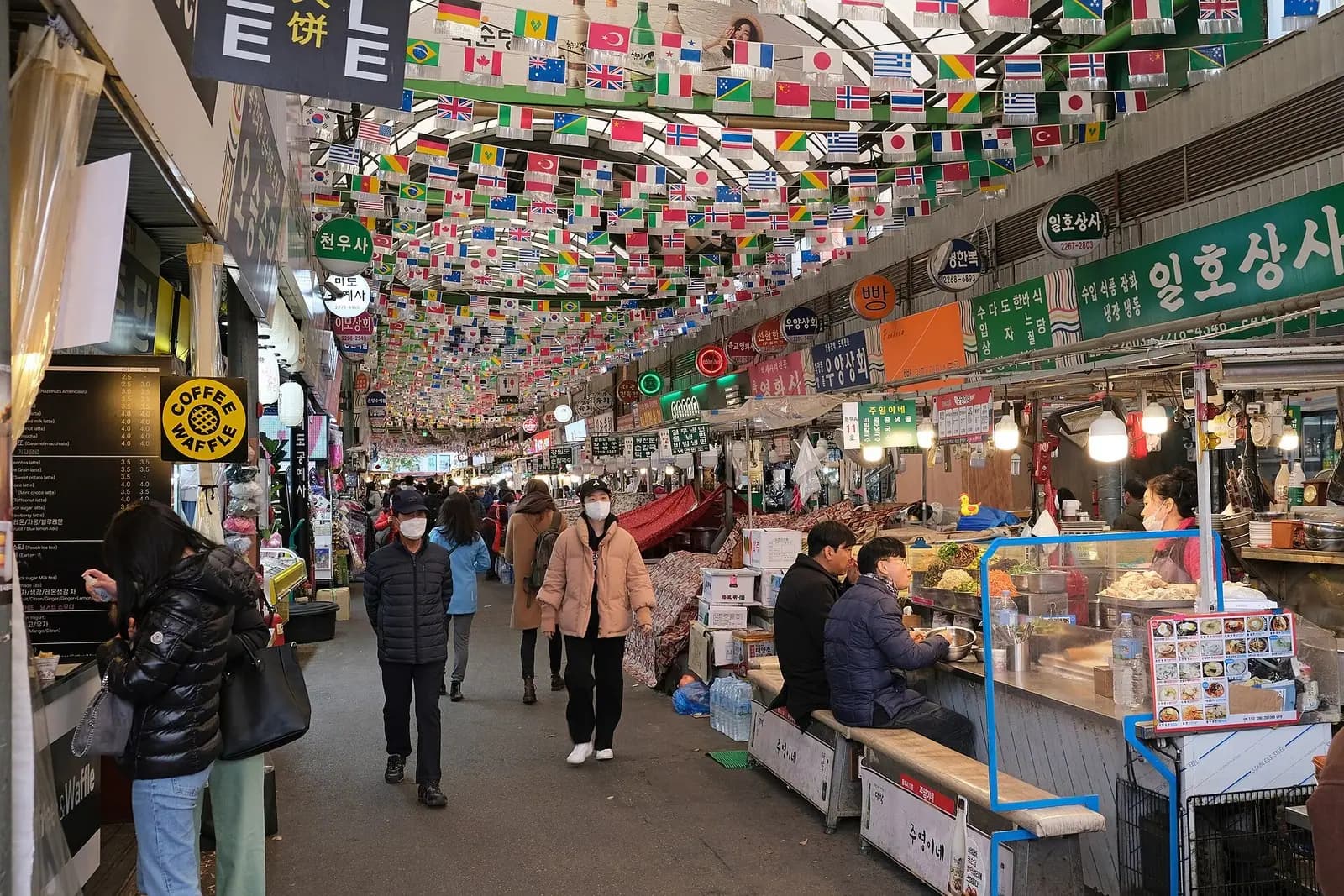 Shoppers walk a covered Gwangjang Market aisle under rows of small national flags