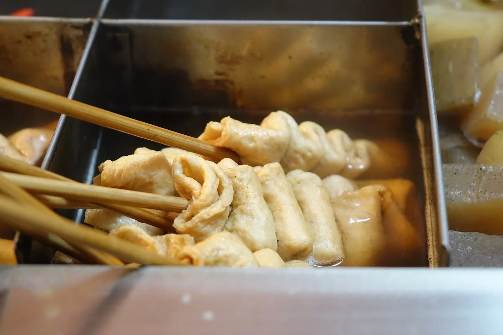 熱いだしの入った金属のトレーに並ぶ、折りたたんで串に刺したオムク