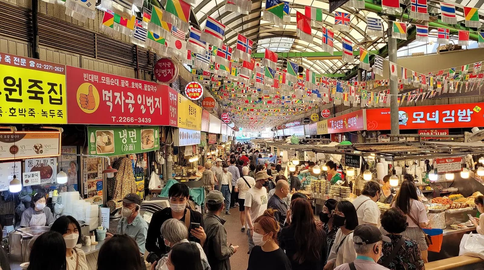 旗と屋台の看板が吊り下がる、ソウルの広蔵市場の混み合った屋根付きの食べ物横丁
