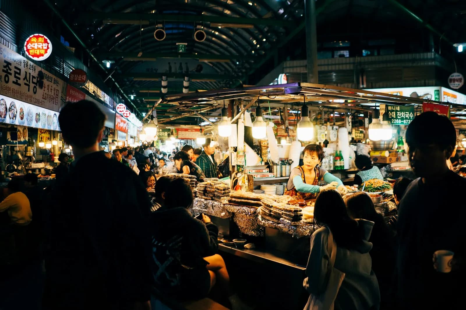 Food stalls under hanging lamps in a covered Korean market, with diners seen from behind