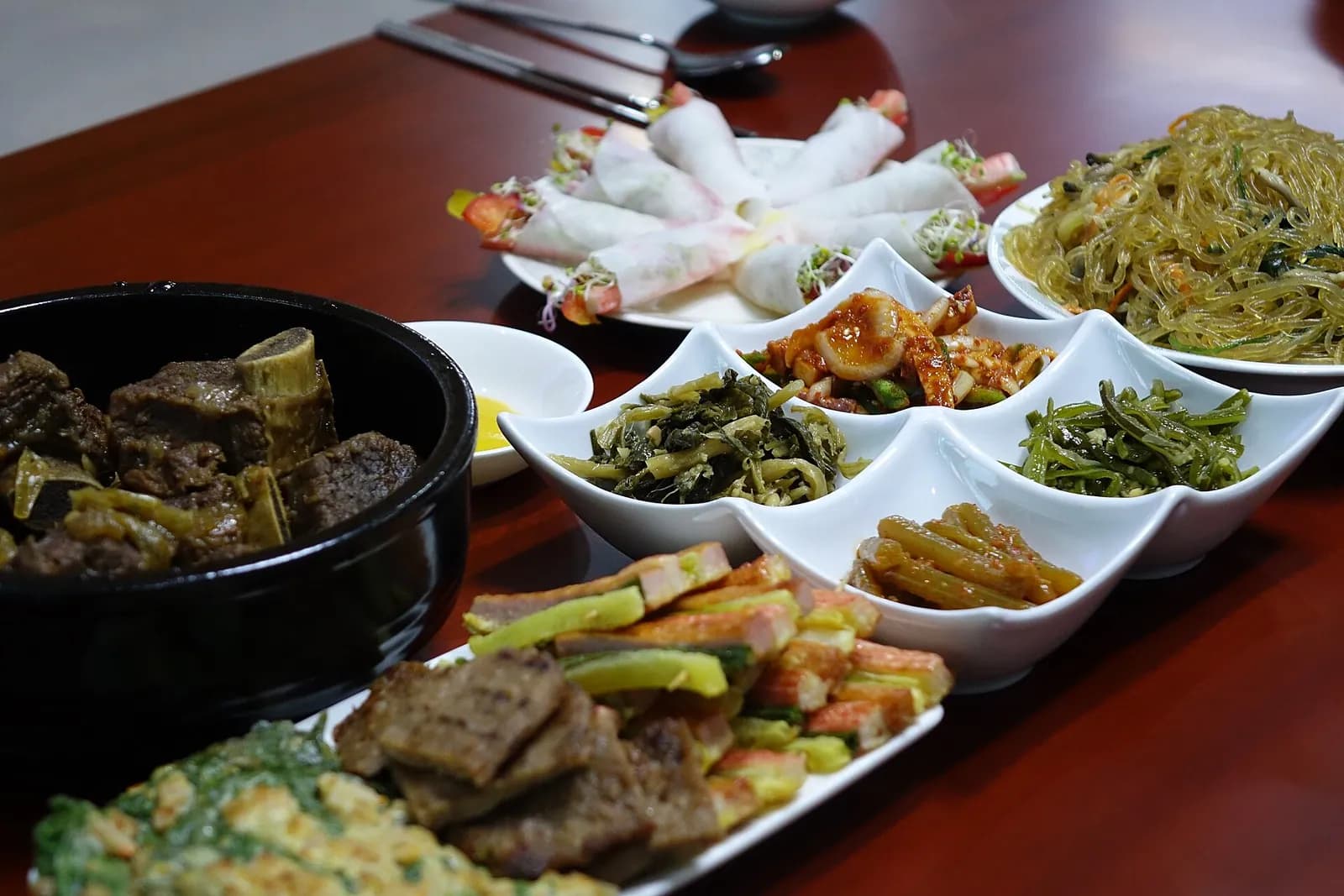 Korean restaurant table with shared plates of japchae, braised ribs, pan-fried jeon and vegetable side dishes
