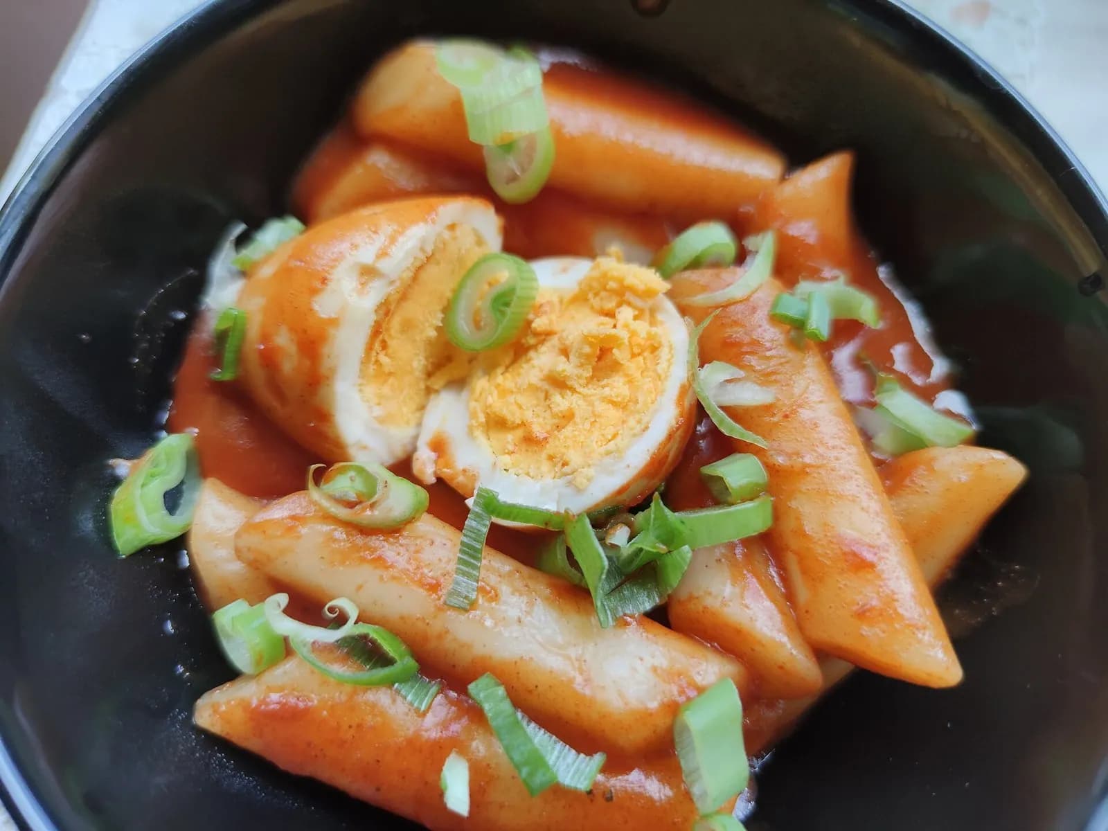 Bowl of tteokbokki rice cakes in red sauce topped with boiled egg and green onion