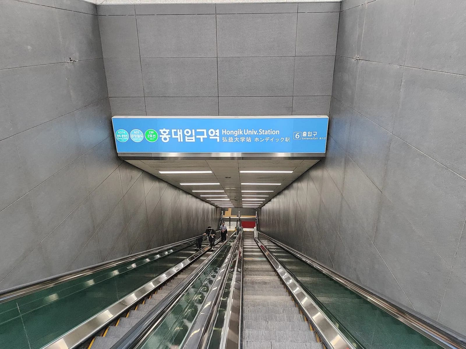 Blue sign for exit 6 of Hongik University Station above a long escalator in Seoul